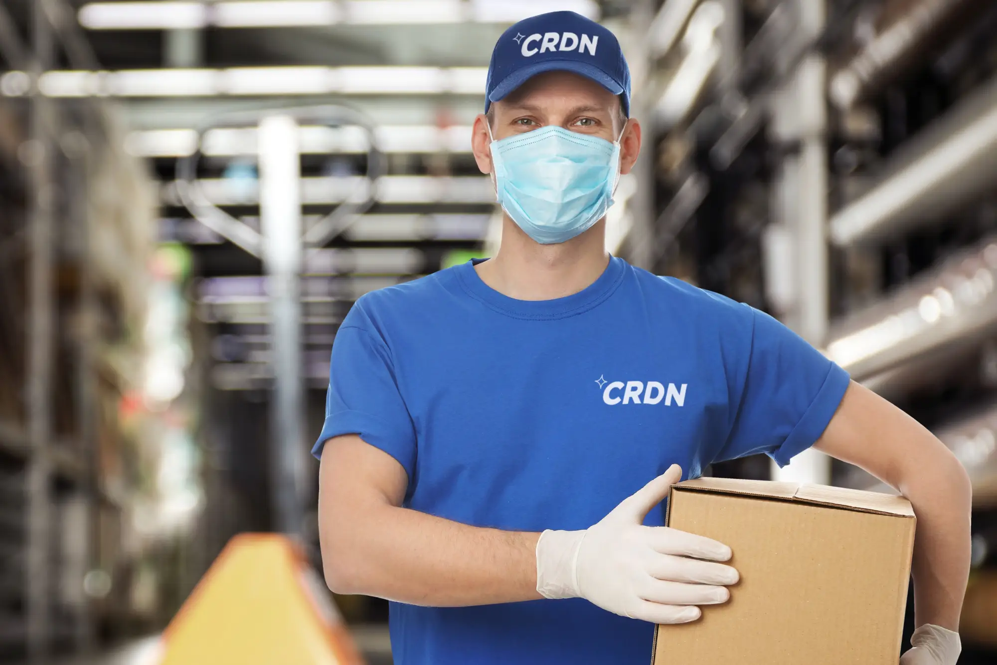 A worker in a blue CRDN shirt and cap, wearing gloves, holds a cardboard box in a warehouse setting