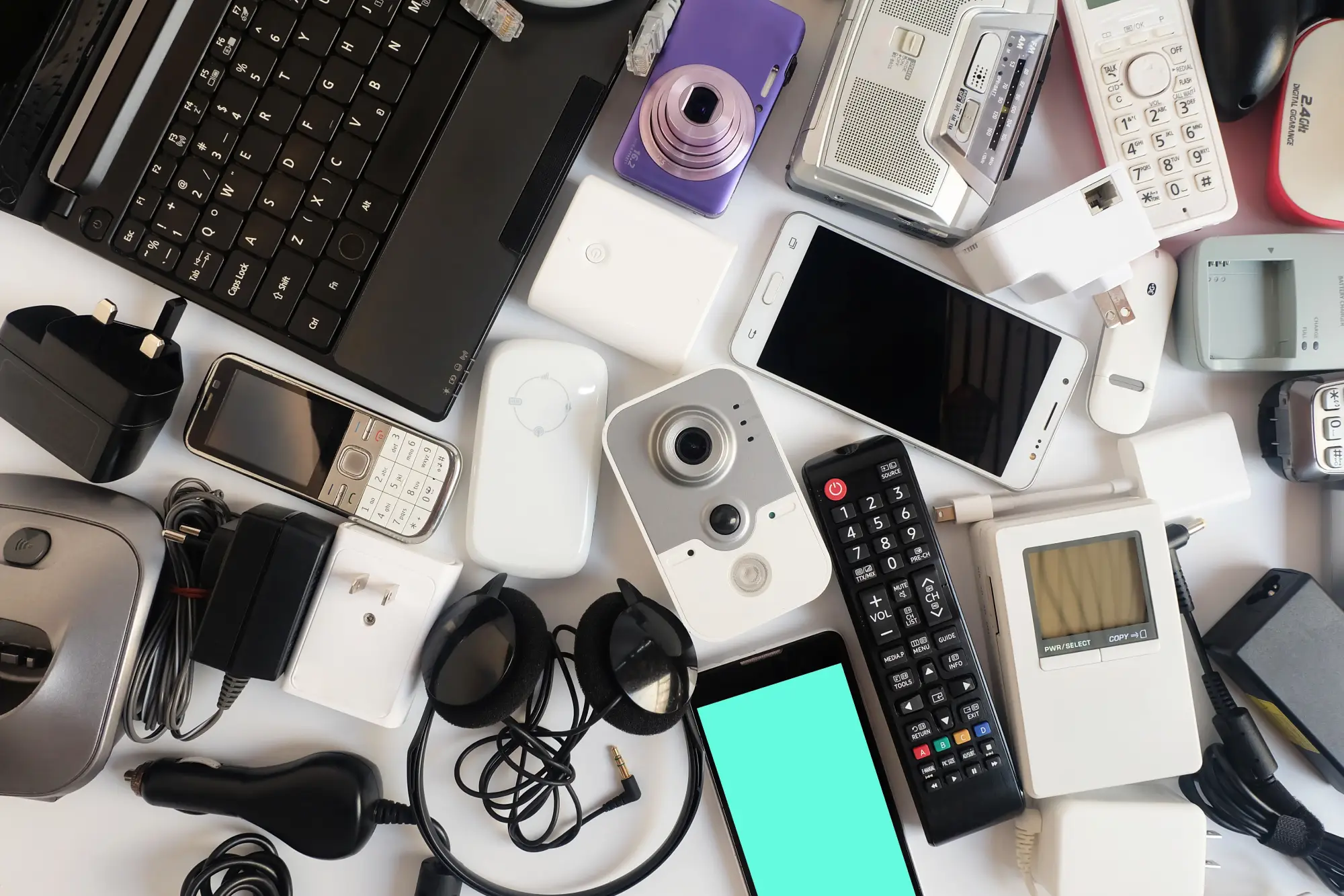 An array of electronic devices and accessories, including a laptop, phones, cameras, chargers, and headphones, scattered on a surface
